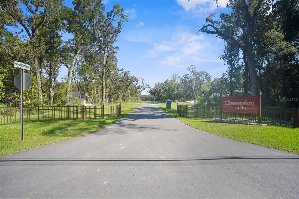 An asphalt road leads into the gated entrance of Champion Estates, flanked by trees — REPUBLICAN (LOT 7) WAY, BROOKSVILLE, FL