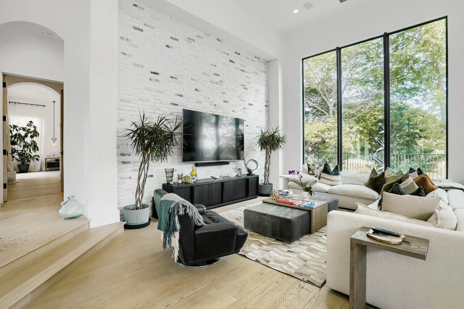 A modern living room features a white brick accent wall, large windows, and neutral furniture — 6179 Vía Venetia S, Delray Beach FL 33484, DELRAY BEACH, FL