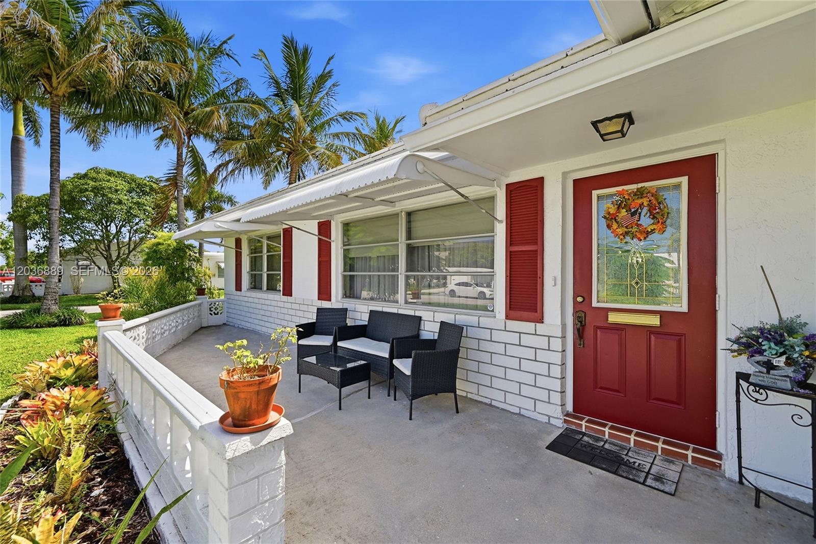 A white brick house exterior features a red front door, patio furniture, and palm trees — 1702 SW 22nd St, Boynton Beach FL 33426, BOYNTON BEACH, FL