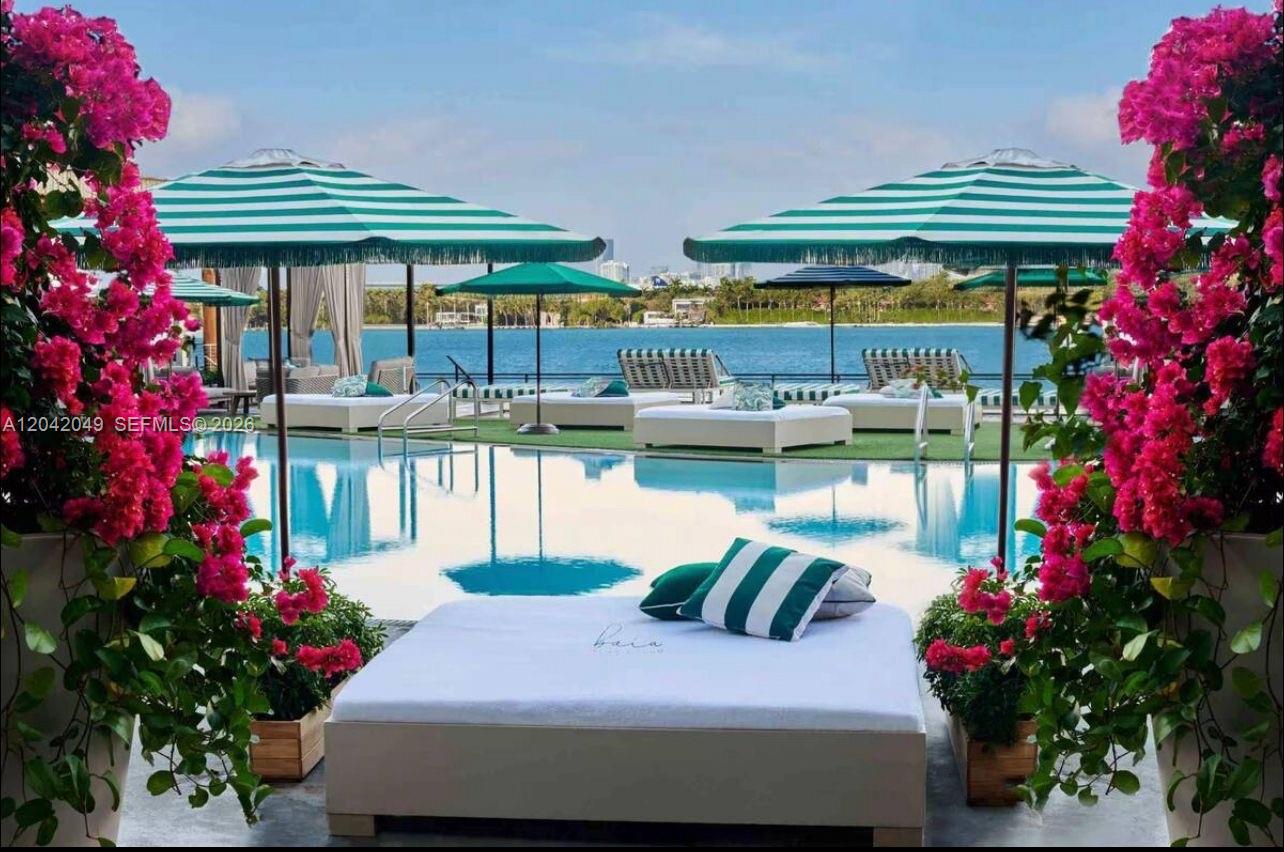 A swimming pool area features lounge chairs, striped umbrellas, and vibrant pink bougainvillea flowers — 1100 West Ave # 1007, Miami Beach FL 33139, MIAMI BEACH, FL