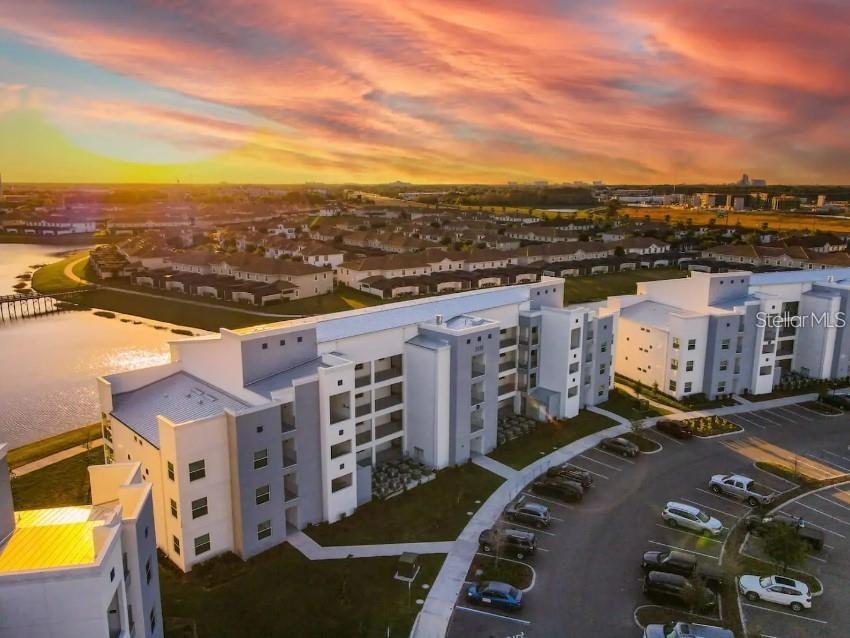 An aerial view shows a modern apartment complex next to a lake at sunset — 4741 CLOCK TOWER DRIVE UNIT 108, KISSIMMEE, FL