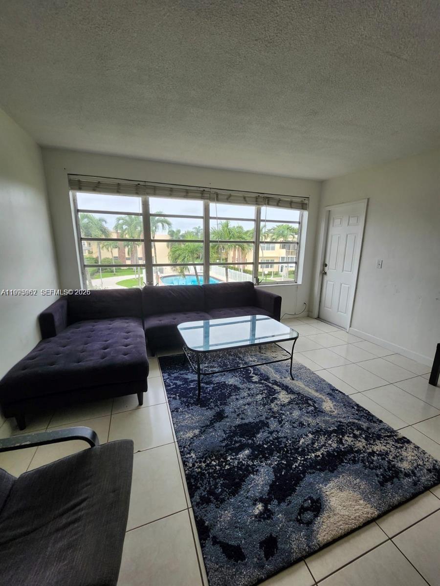 A living room features a dark sectional sofa, glass coffee table, tile flooring, and windows — 851 NE 14th Ave # 420, Hallandale Beach FL 33009, HALLANDALE BEACH, FL