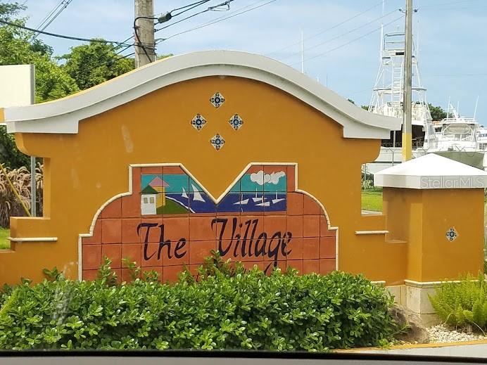 A yellow stucco sign for The Village stands in front of a marina with boats — The Village at the Hill MANGO BEACH CORNER UNIT 116, CEIBA, PR