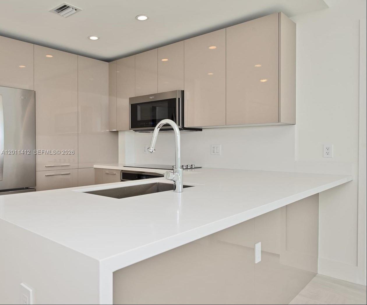 A modern kitchen features glossy beige cabinetry, white countertops, and stainless steel appliances — 2000 Metropica Way # 2206, Sunrise FL 33323, SUNRISE, FL