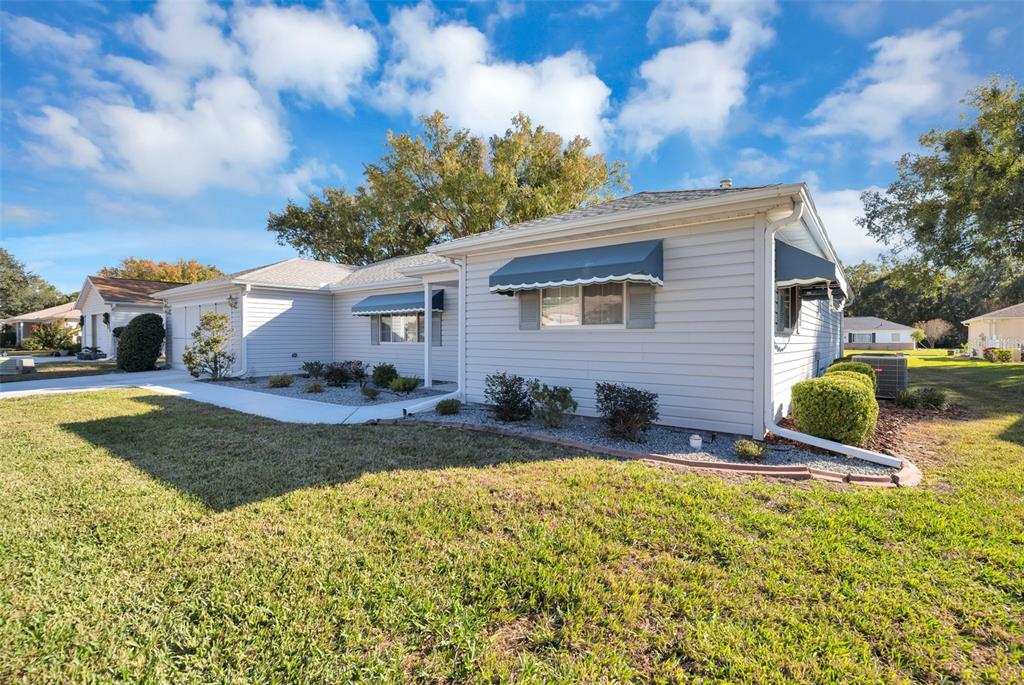 A light-colored, single-story house with blue window awnings and a landscaped front yard — 9921 SE 138TH LOOP, SUMMERFIELD, FL