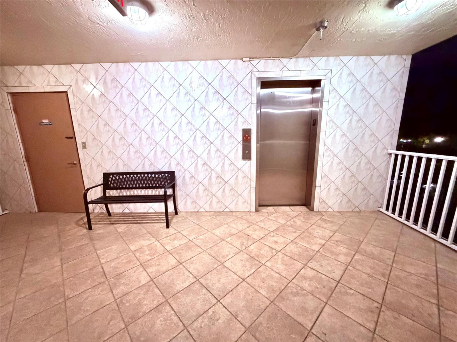 An outdoor elevator lobby features tiled walls and flooring, a metal bench, and a door — 7302 S Devon Drive # 307, Tamarac FL 33321, TAMARAC, FL