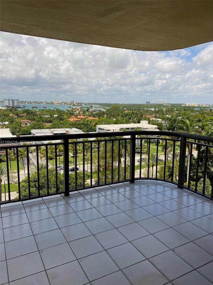 A tiled balcony with a black metal railing overlooks a tropical landscape and distant water — 10175 Collins Ave # 807, Bal Harbour FL 33154, BAL HARBOUR, FL