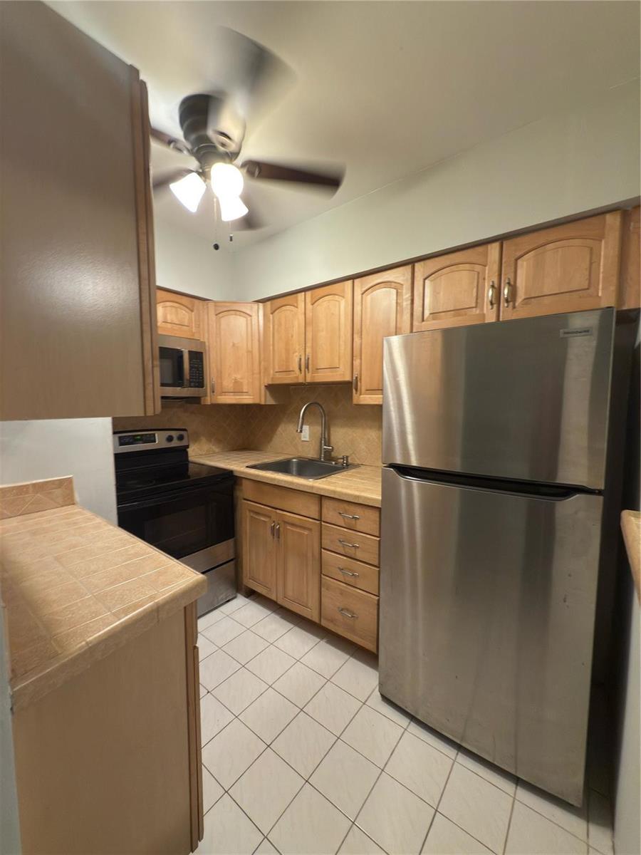 A small kitchen features light wood cabinets, tile countertops, stainless steel appliances, and tile flooring — 600 Layne Boulevard # 117, Hallandale Beach FL 33009, HALLANDALE BEACH, FL