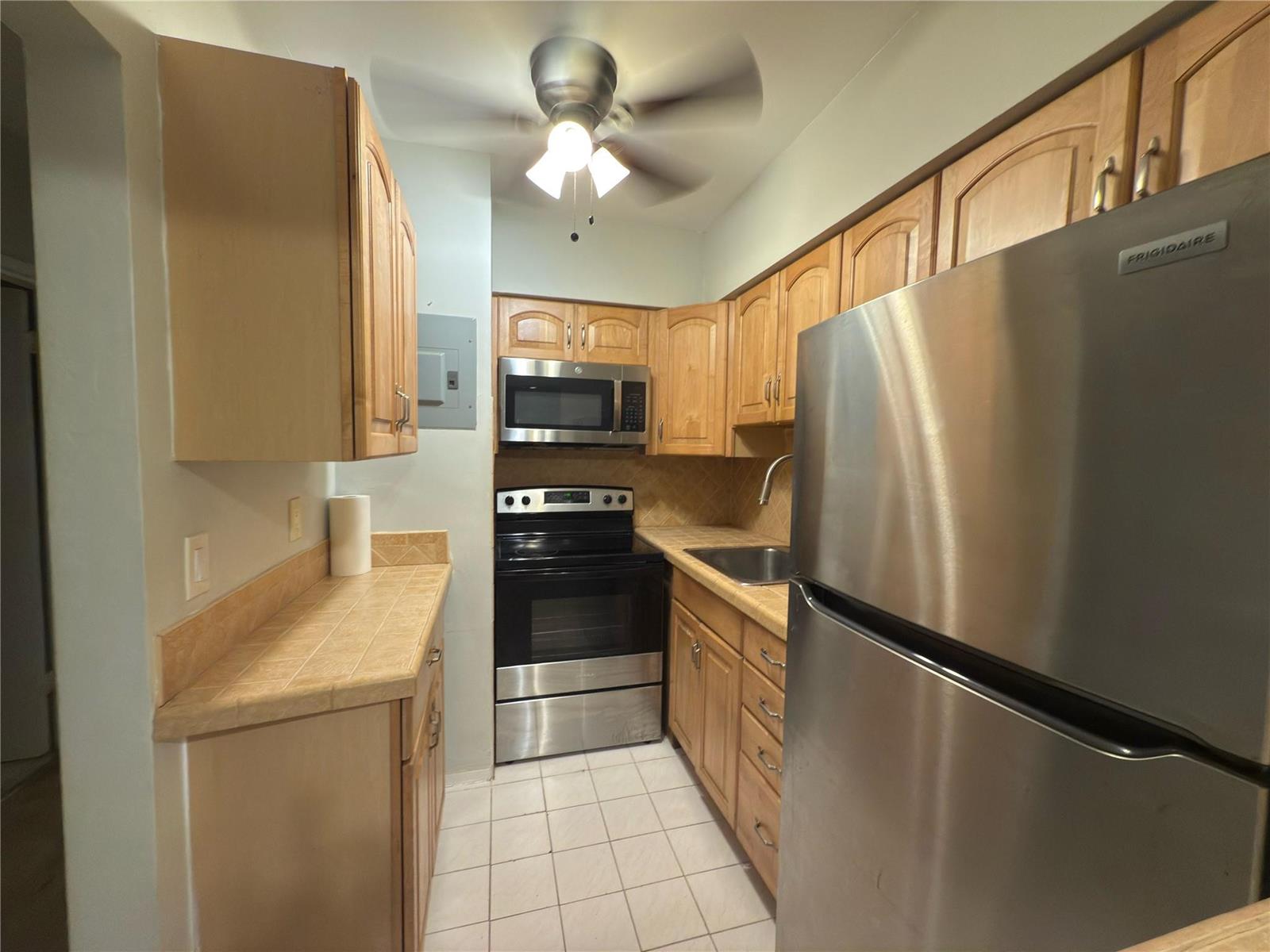 A galley kitchen features light wood cabinets, stainless steel appliances, and tan tile countertops — 600 Layne Boulevard # 117, Hallandale Beach FL 33009, HALLANDALE BEACH, FL