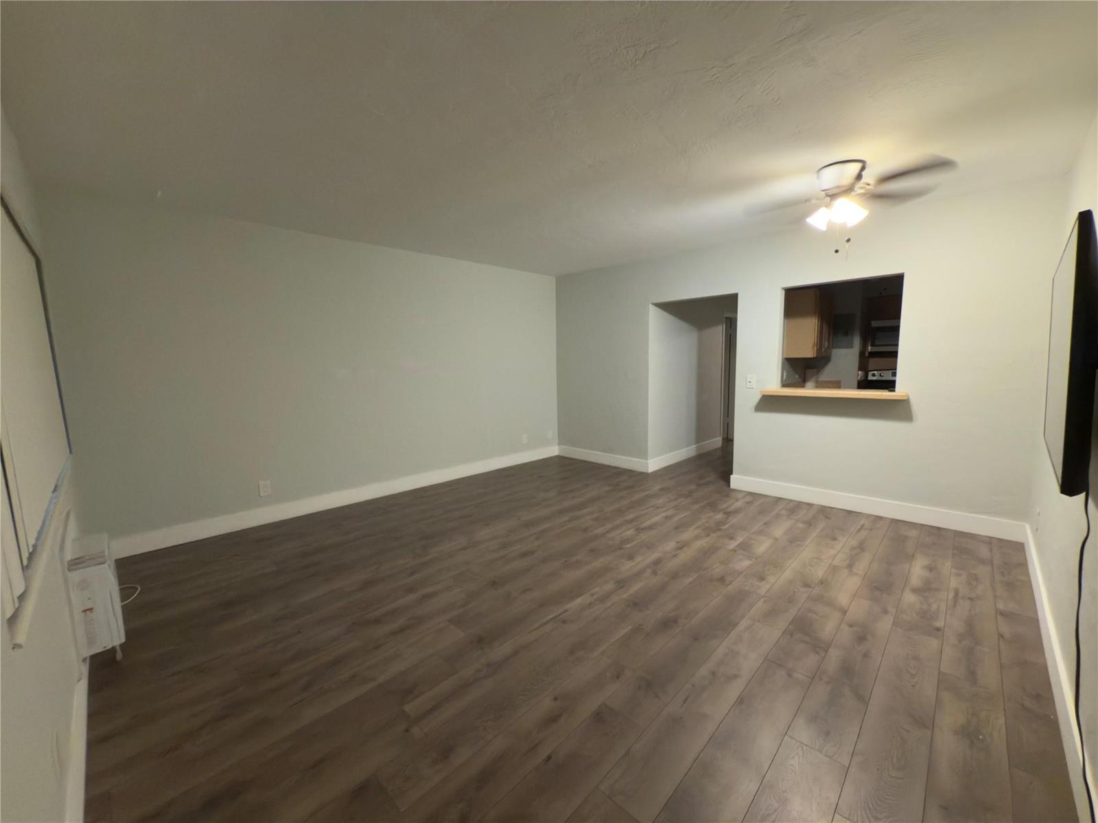An empty living room features light gray walls, dark wood-look flooring, and a kitchen pass-through — 600 Layne Boulevard # 117, Hallandale Beach FL 33009, HALLANDALE BEACH, FL