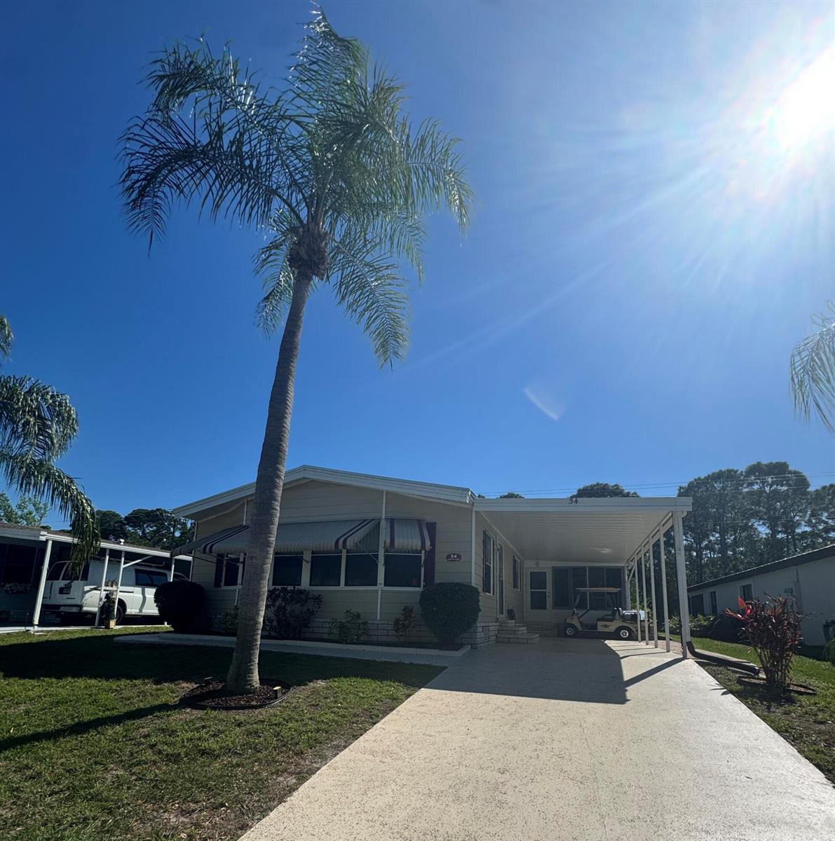 A single-story mobile home with a palm tree, carport, and driveway under a sunny sky — 54 Calle De Lagos, Fort Pierce FL 34951, FORT PIERCE, FL