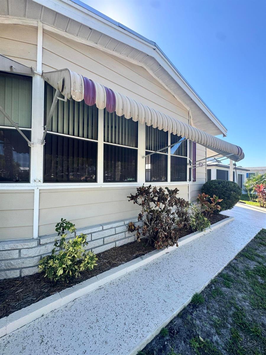The exterior of a mobile home features beige siding, a striped awning, and landscaping — 54 Calle De Lagos, Fort Pierce FL 34951, FORT PIERCE, FL