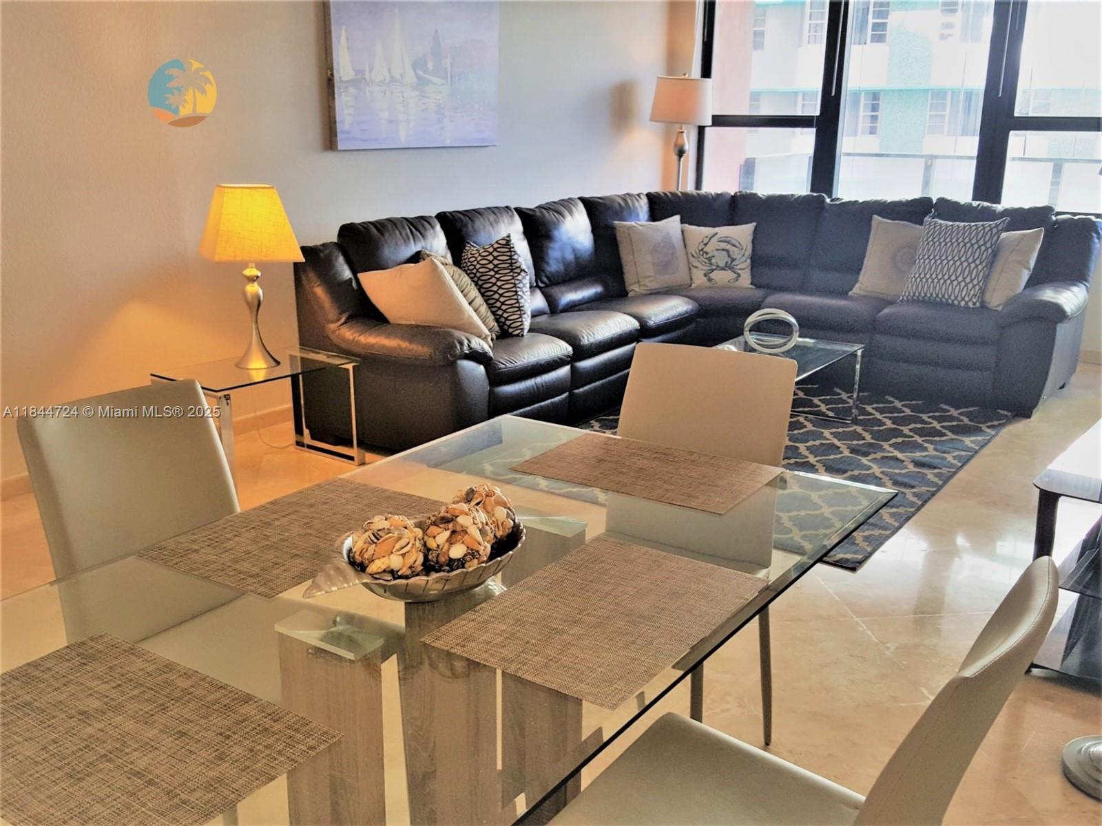 A dining area with a glass table sits in front of a large leather sectional sofa — 5225 Collins Ave # 510, Miami Beach FL 33140, MIAMI BEACH, FL