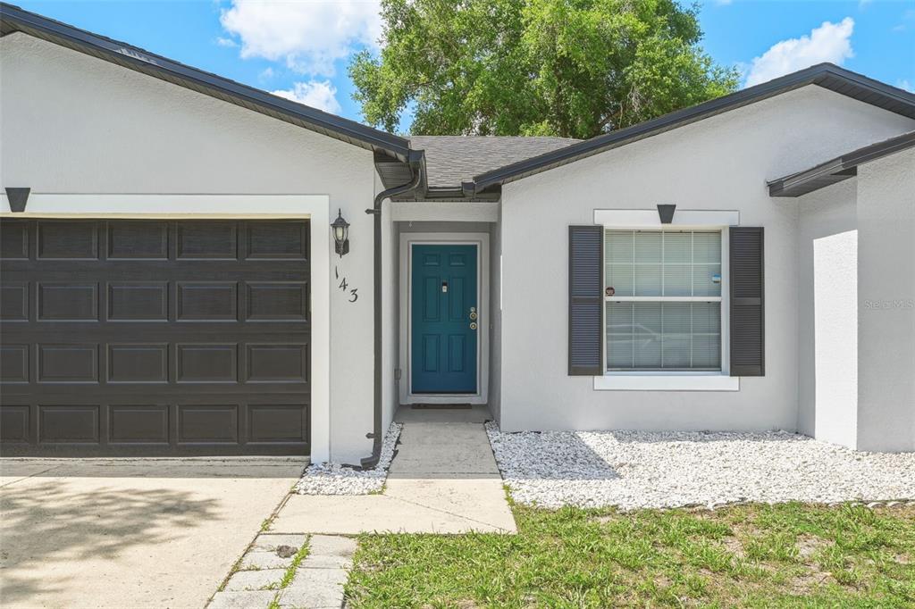 A single-story house exterior features a teal front door, a garage, and a gravel yard — 143 LAUREL RIDGE AVENUE, OCOEE, FL