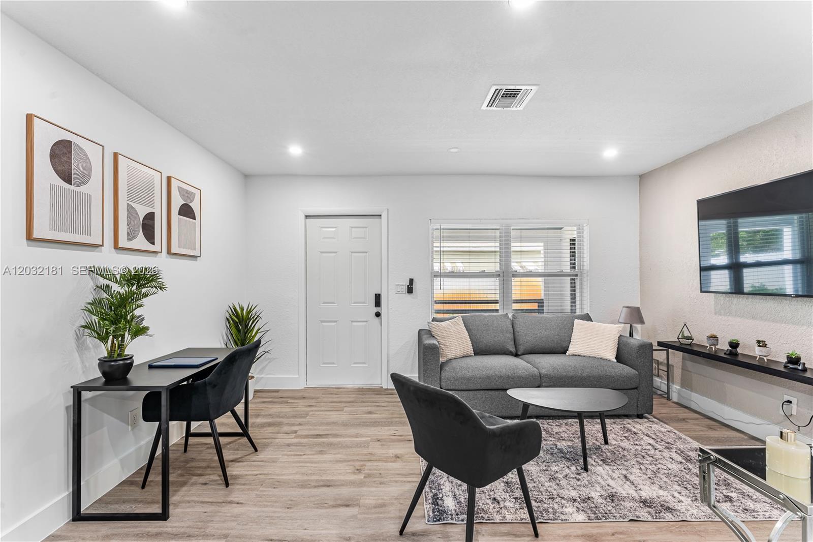 A living room features a gray sofa, a desk with a chair, and a wall-mounted television — 1521 NE 118th Ter # 1, Miami FL 33161, MIAMI, FL