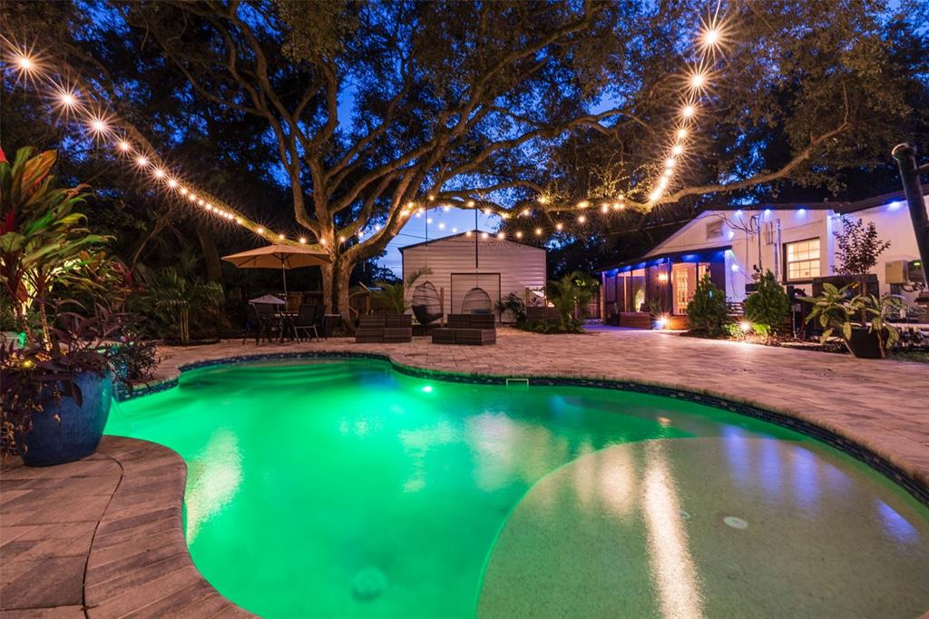 A backyard swimming pool with green lighting is surrounded by a stone patio and string lights — 8701 64TH STREET N, PINELLAS PARK, FL