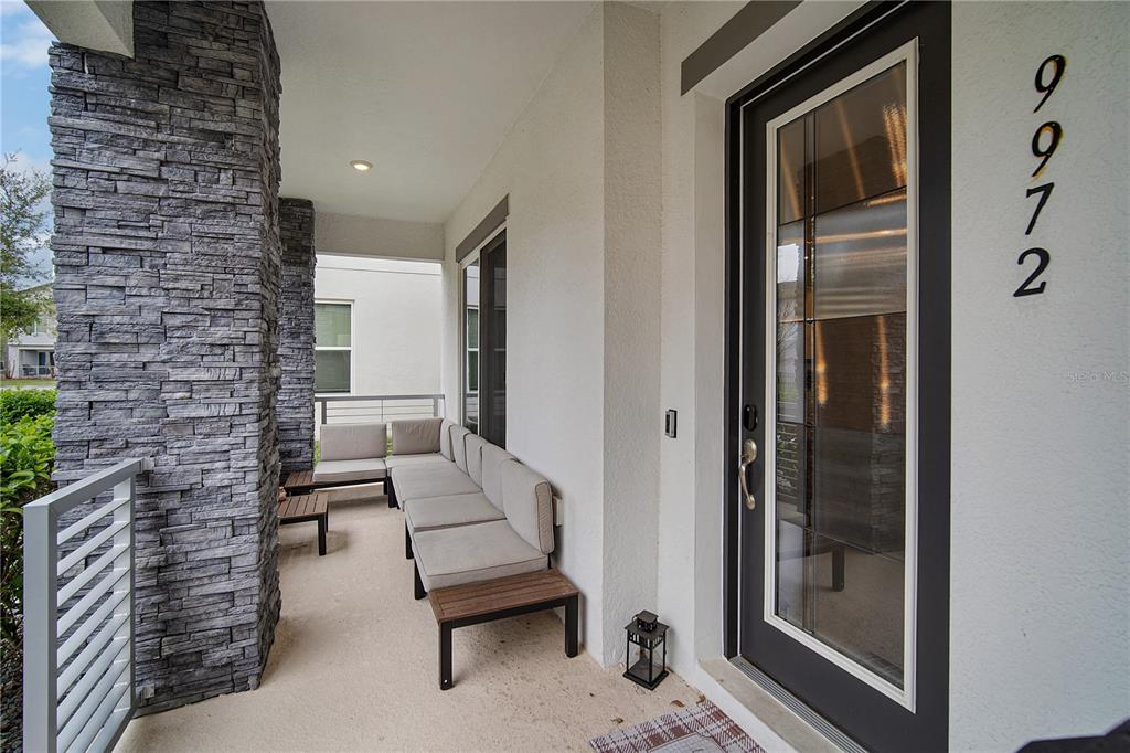A covered porch features stone pillars, light-colored flooring, modular seating, and a glass-paneled front door — 9972 PALLIDA HICKORY WAY, WINTER GARDEN, FL