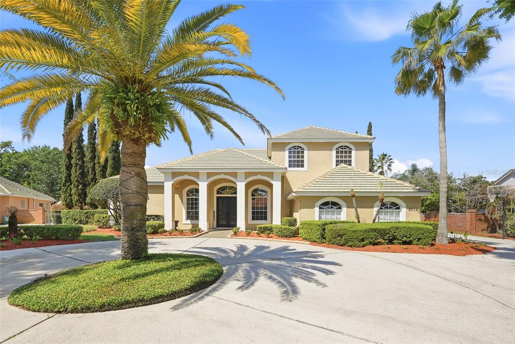 A two-story stucco house with a circular driveway and palm trees under a blue sky — 9119 BALMORAL MEWS SQ, WINDERMERE, FL