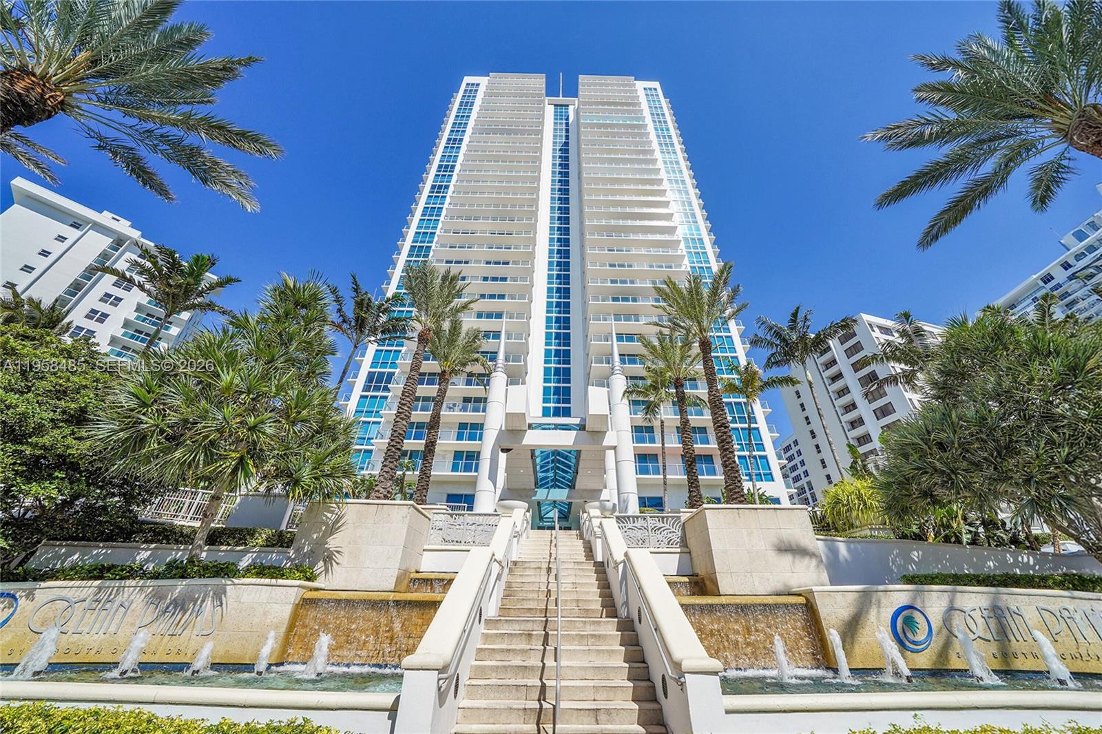 A tall residential building features a central staircase with fountains and surrounding palm trees — 3101 S OCEAN # 1802, Hollywood FL 33019, HOLLYWOOD, FL