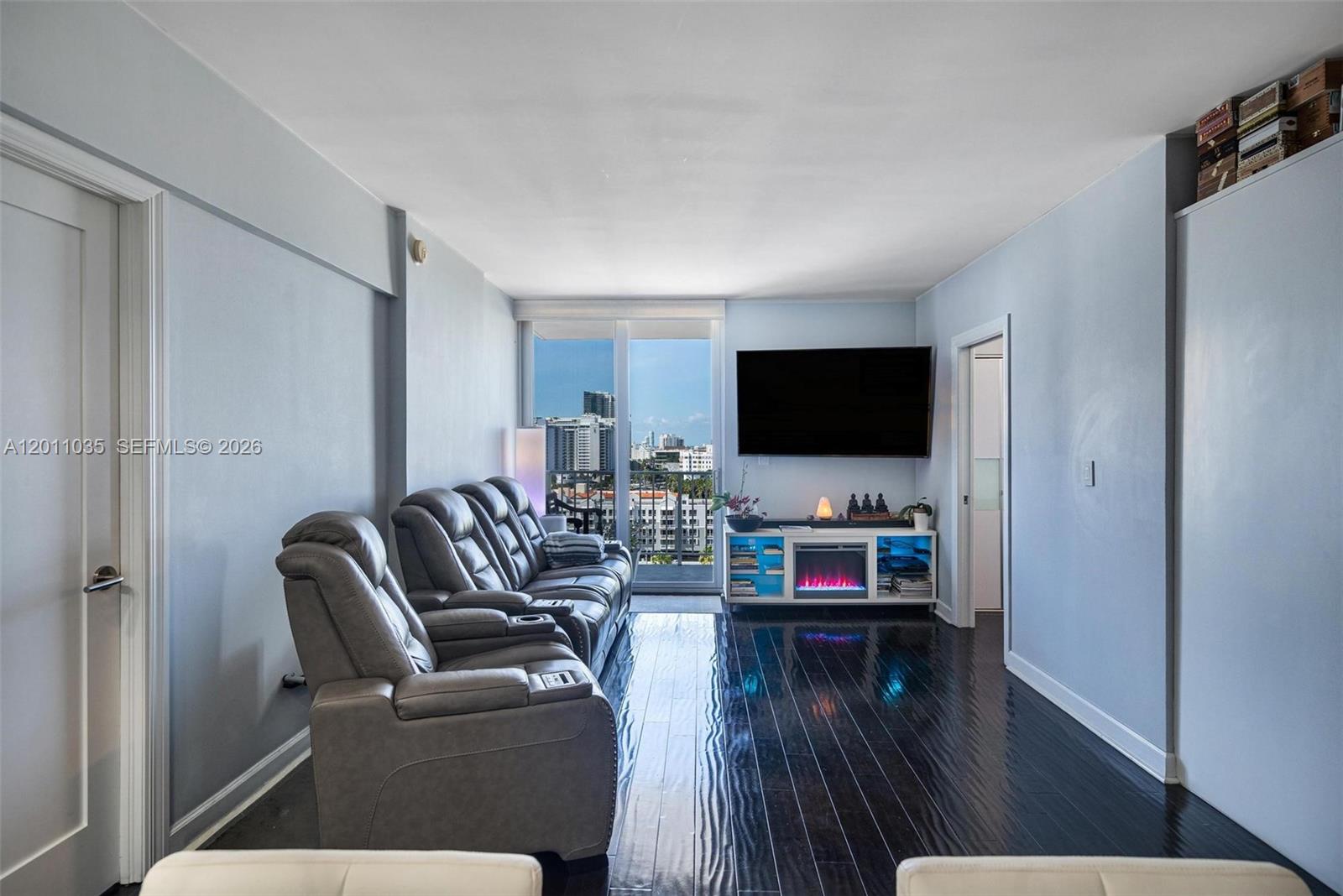 A living room features dark wood floors, gray reclining sofas, and a wall-mounted television — 2829 Indian Creek Dr # 1209, Miami Beach FL 33140, MIAMI BEACH, FL