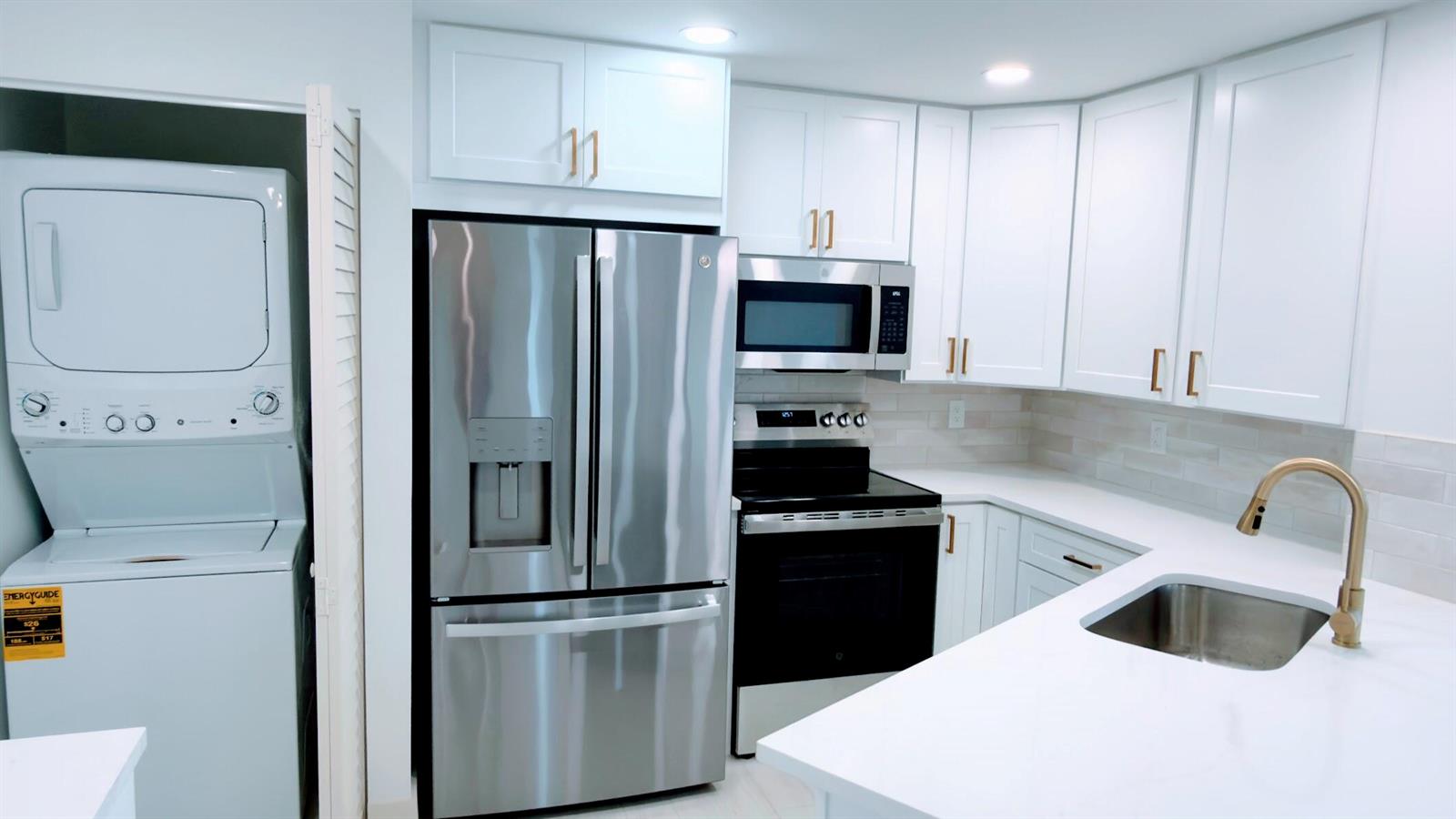 A modern kitchen features white cabinets, stainless steel appliances, and a stacked washer and dryer — 7547 S Oriole Boulevard # 101, Delray Beach FL 33446, DELRAY BEACH, FL