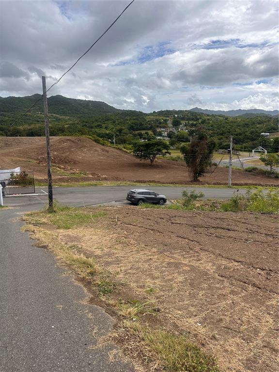 A view of a cleared, sloped dirt lot with a car parked on the road below — BO. SAN IDELFONSO, COAMO, PR