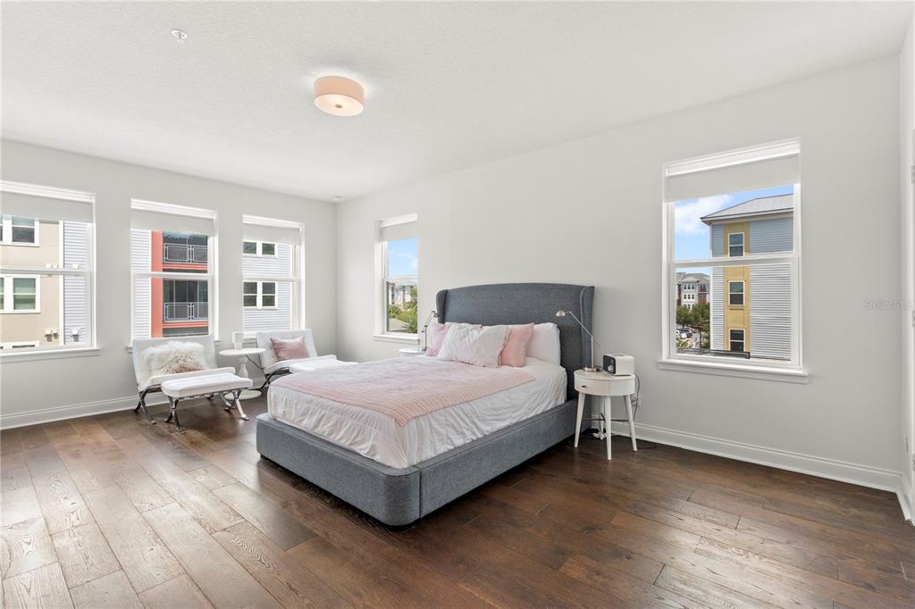 A bedroom with hardwood floors, a gray upholstered bed, and large windows overlooking neighboring buildings — 7573 LAUREATE BOULEVARD UNIT 1-307, ORLANDO, FL