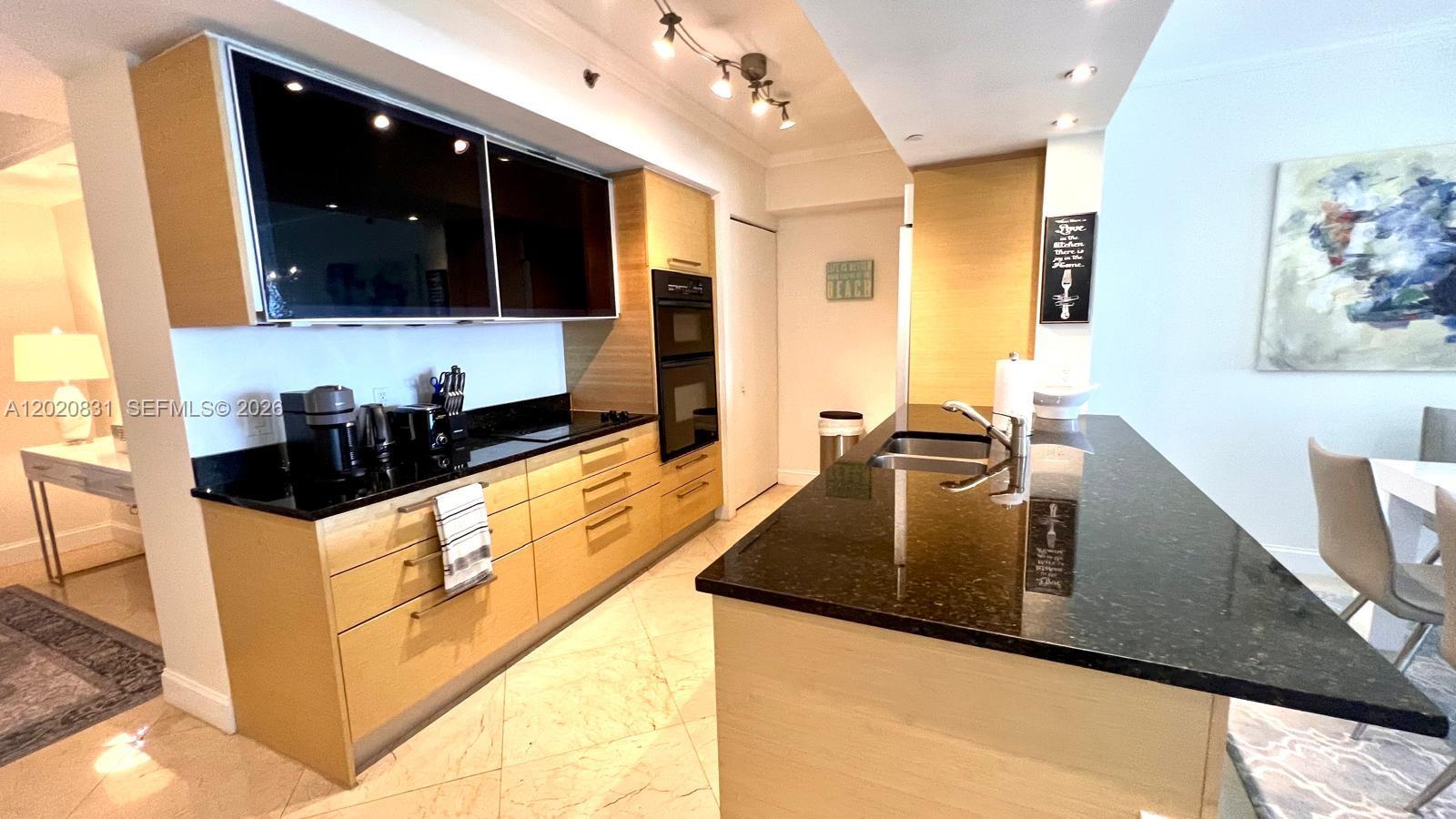 A modern kitchen features light wood cabinets, black countertops, and a central island with sink — 1830 S Ocean Dr # 2204, Hallandale Beach FL 33009, HALLANDALE BEACH, FL