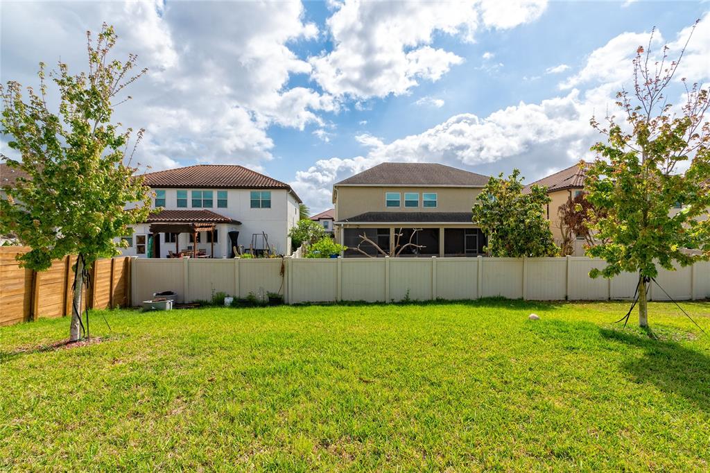 A grassy backyard features two young trees and a tan vinyl fence against three houses — 14815 MILFOIL AVENUE, ORLANDO, FL