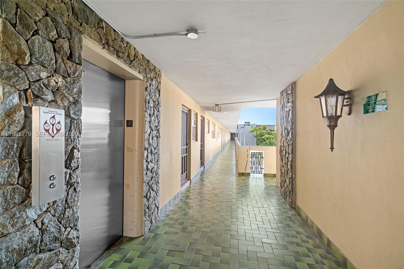 An outdoor apartment hallway features stone-accented walls, green tiled flooring, and an elevator entrance — 1690 NE 191st St # 1, Miami FL 33179, MIAMI, FL