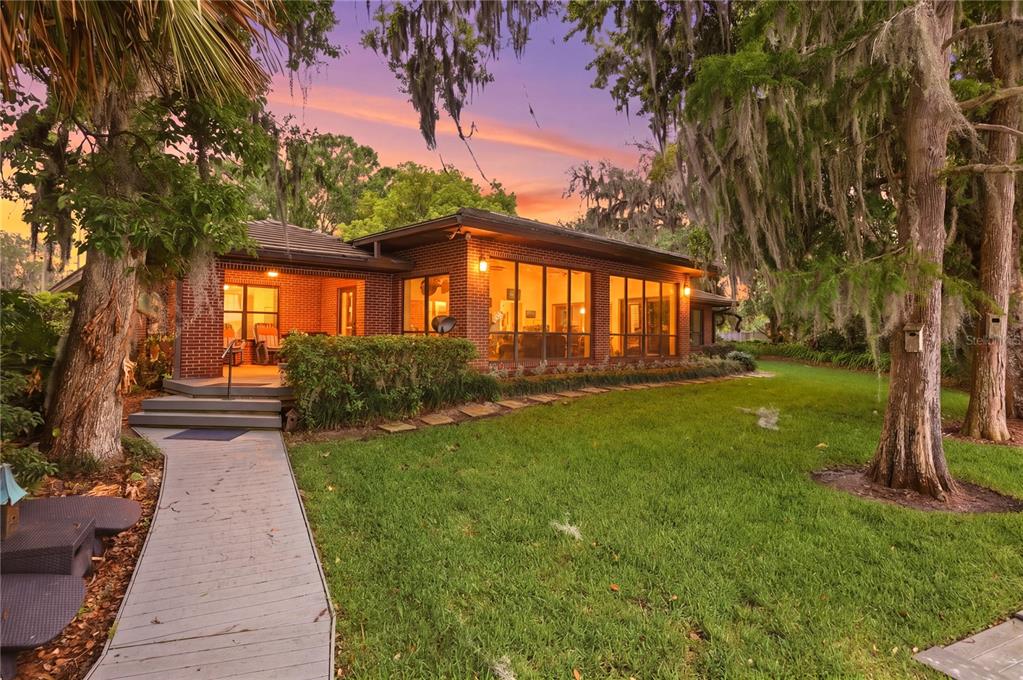 A brick house with large windows and a wooden walkway sits on a grassy lawn — 4910 E LAKE DRIVE, WINTER SPRINGS, FL