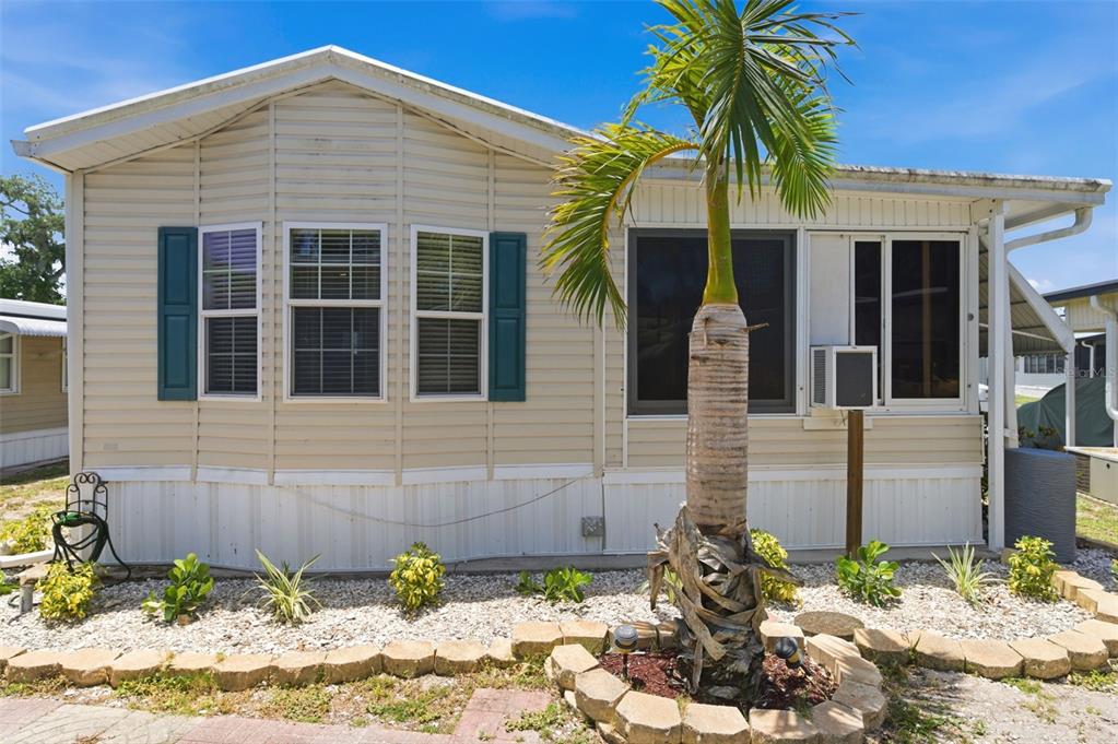 A beige mobile home features teal shutters, a palm tree, and a stone-bordered garden bed — 3275 26TH AVENUE E UNIT 134, BRADENTON, FL