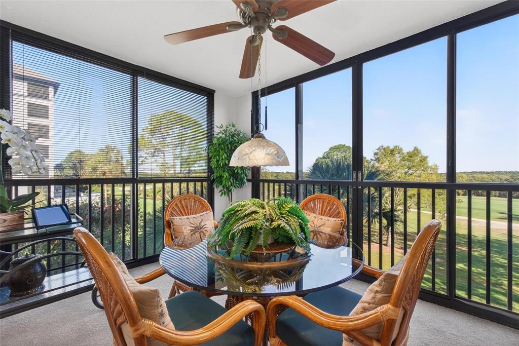 A sunroom features a glass-topped dining table, wicker chairs, and floor-to-ceiling windows overlooking greenery — 14820 RUE DE BAYONNE UNIT 302, CLEARWATER, FL