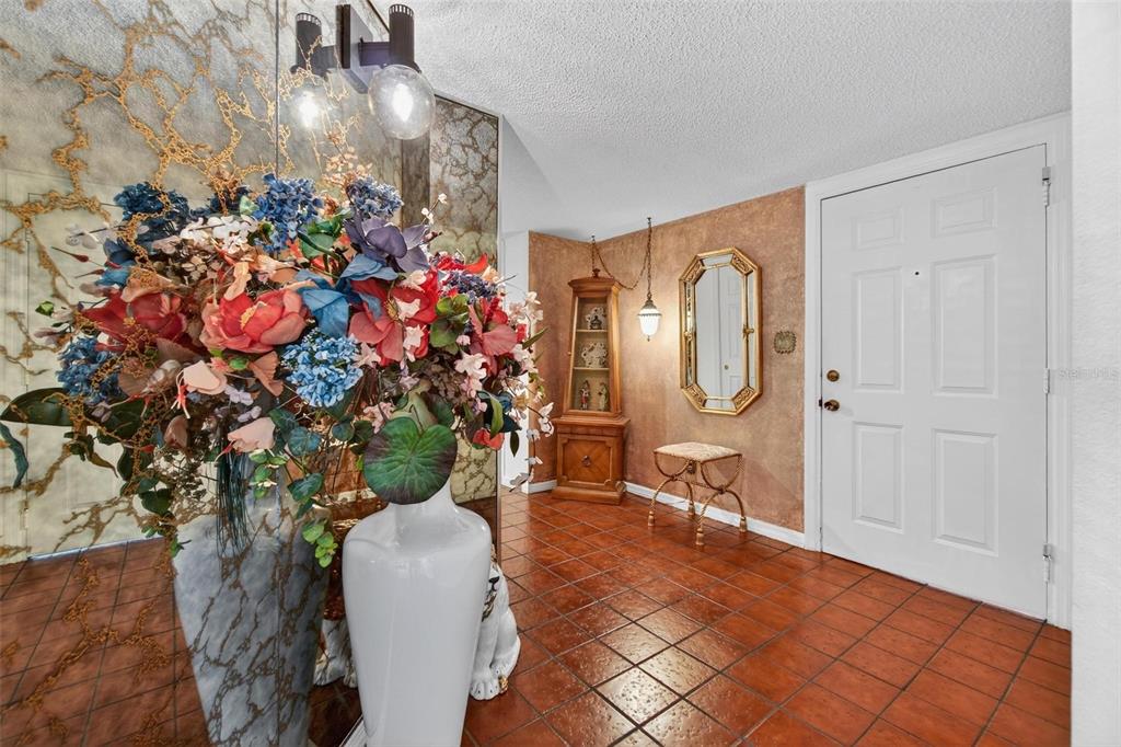An entryway features terracotta tile flooring, a large floral arrangement, and a white front door — 14820 RUE DE BAYONNE UNIT 302, CLEARWATER, FL