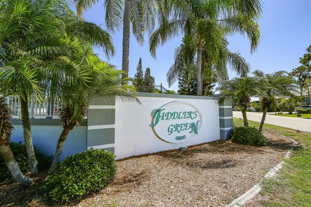 A white stucco monument sign for Fiddler's Green stands outdoors surrounded by palm trees — 6800 PLACIDA ROAD UNIT TH17, ENGLEWOOD, FL