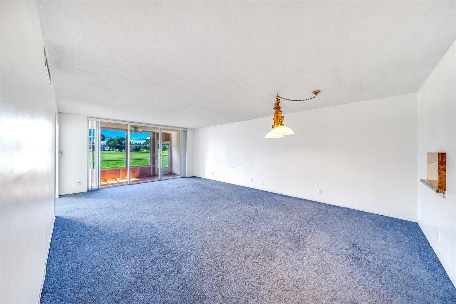 An empty living room features blue carpeting, white walls, a hanging light, and sliding doors — 2601 S Course Drive # 111, Pompano Beach FL 33069, POMPANO BEACH, FL