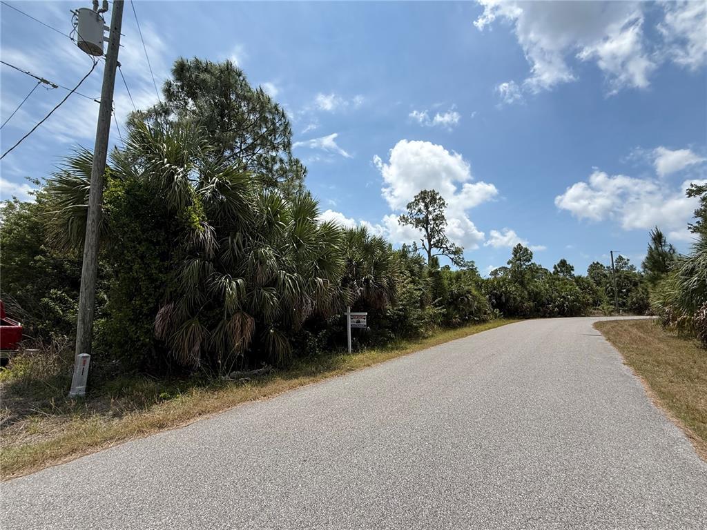 A paved road curves past a wooded lot with a real estate sign — ORCHARD CIRCLE, NORTH PORT, FL