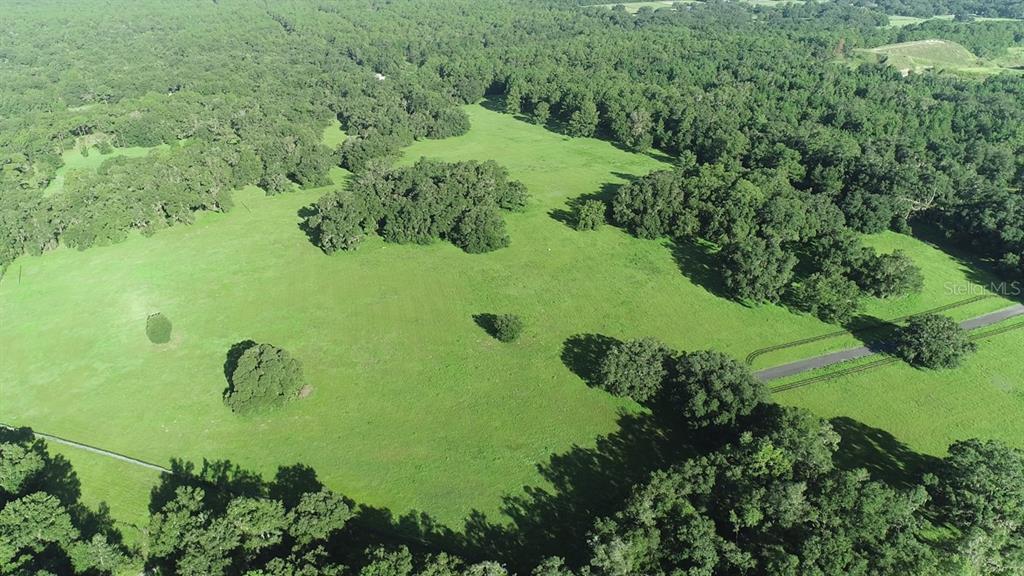 An aerial view shows a large, grassy pasture surrounded by dense woods and a paved road — 1077 SW 144TH COURT ROAD, OCALA, FL