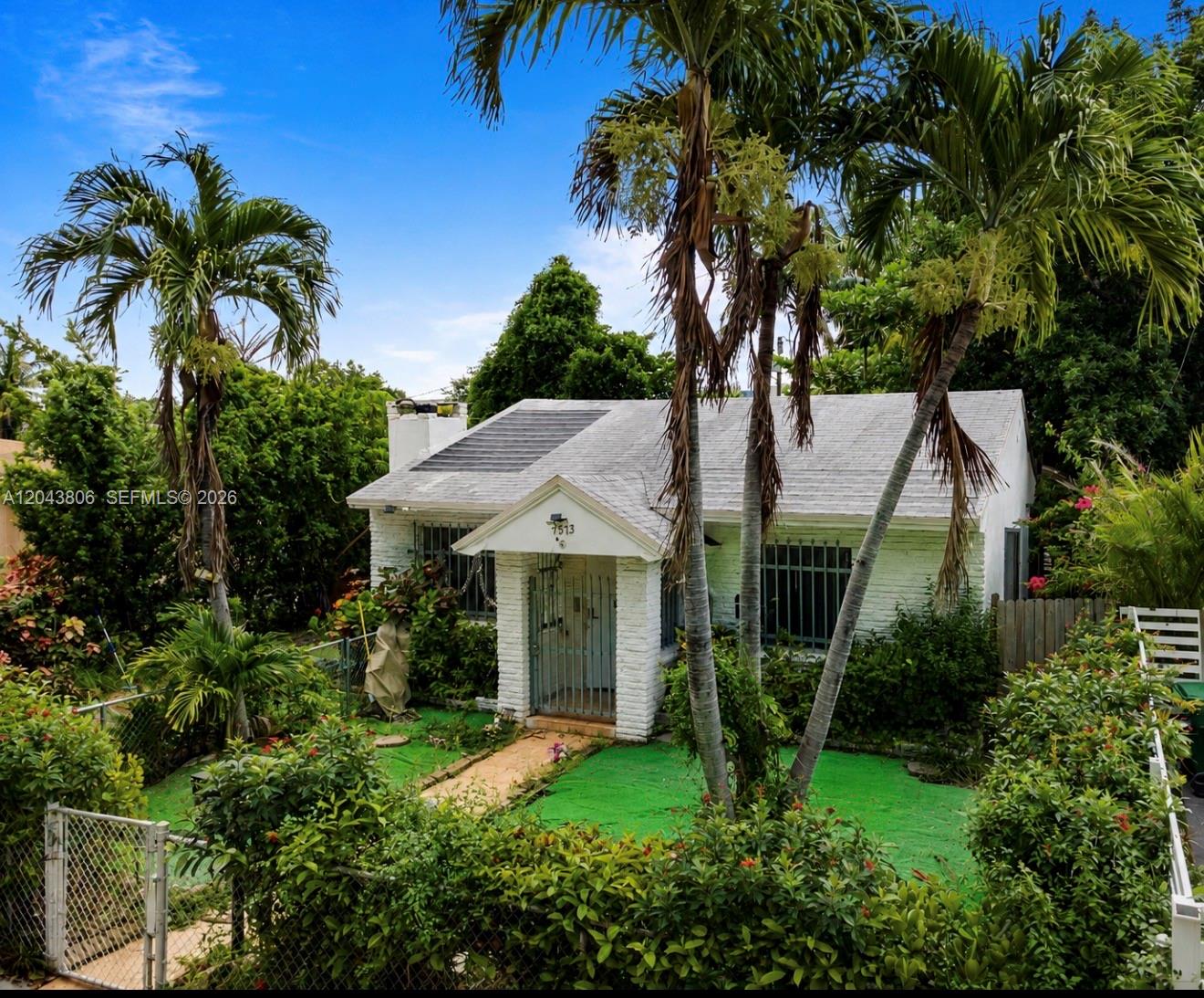 A white, single-story house with a metal-gated entrance is surrounded by lush tropical landscaping — 7573 NW 12th Ct, Miami FL 33147, MIAMI, FL