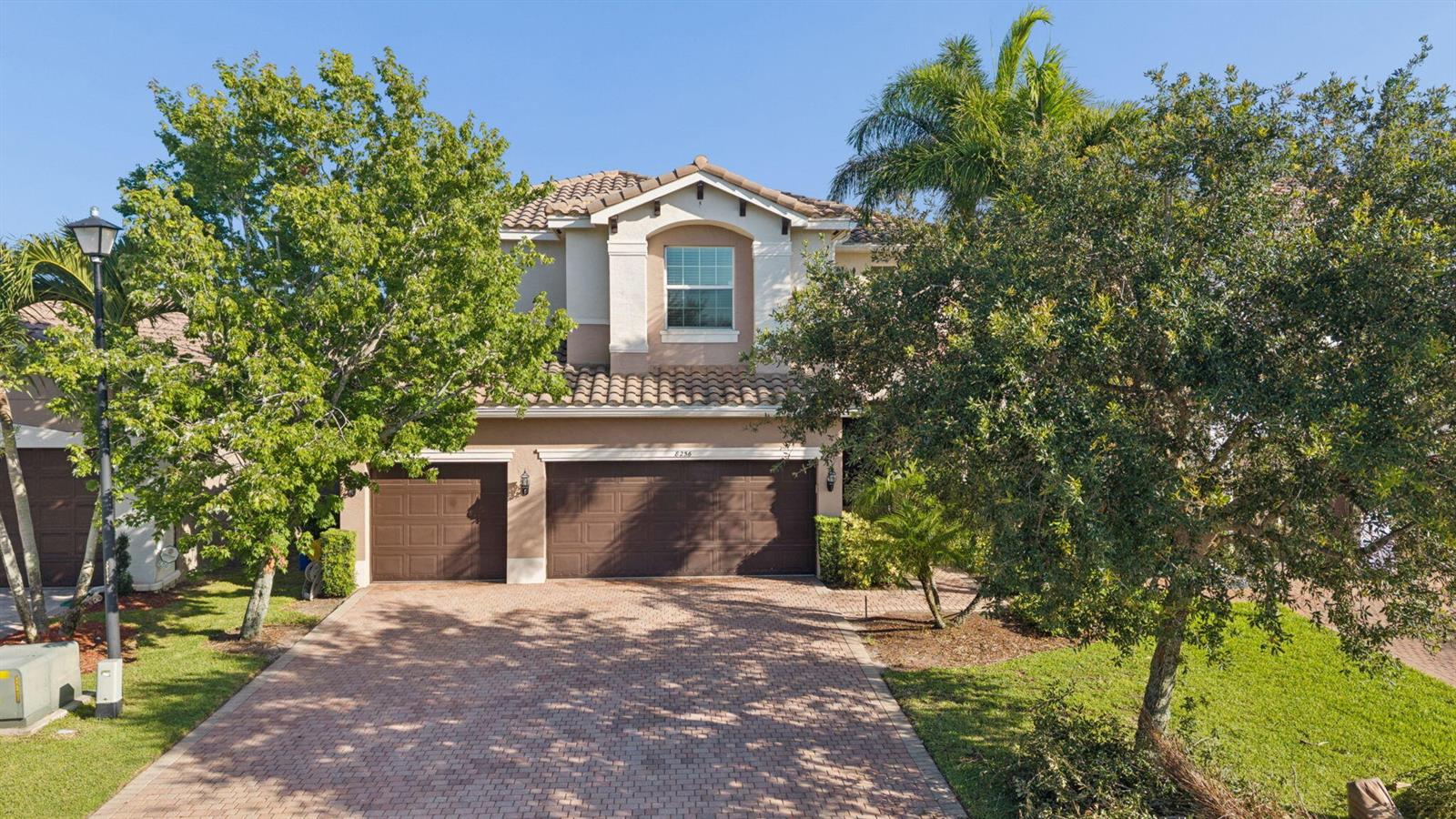A two-story house with a three-car garage and a paved driveway sits behind mature trees — 8256 Savara Streams Lane, Boynton Beach FL 33473, BOYNTON BEACH, FL