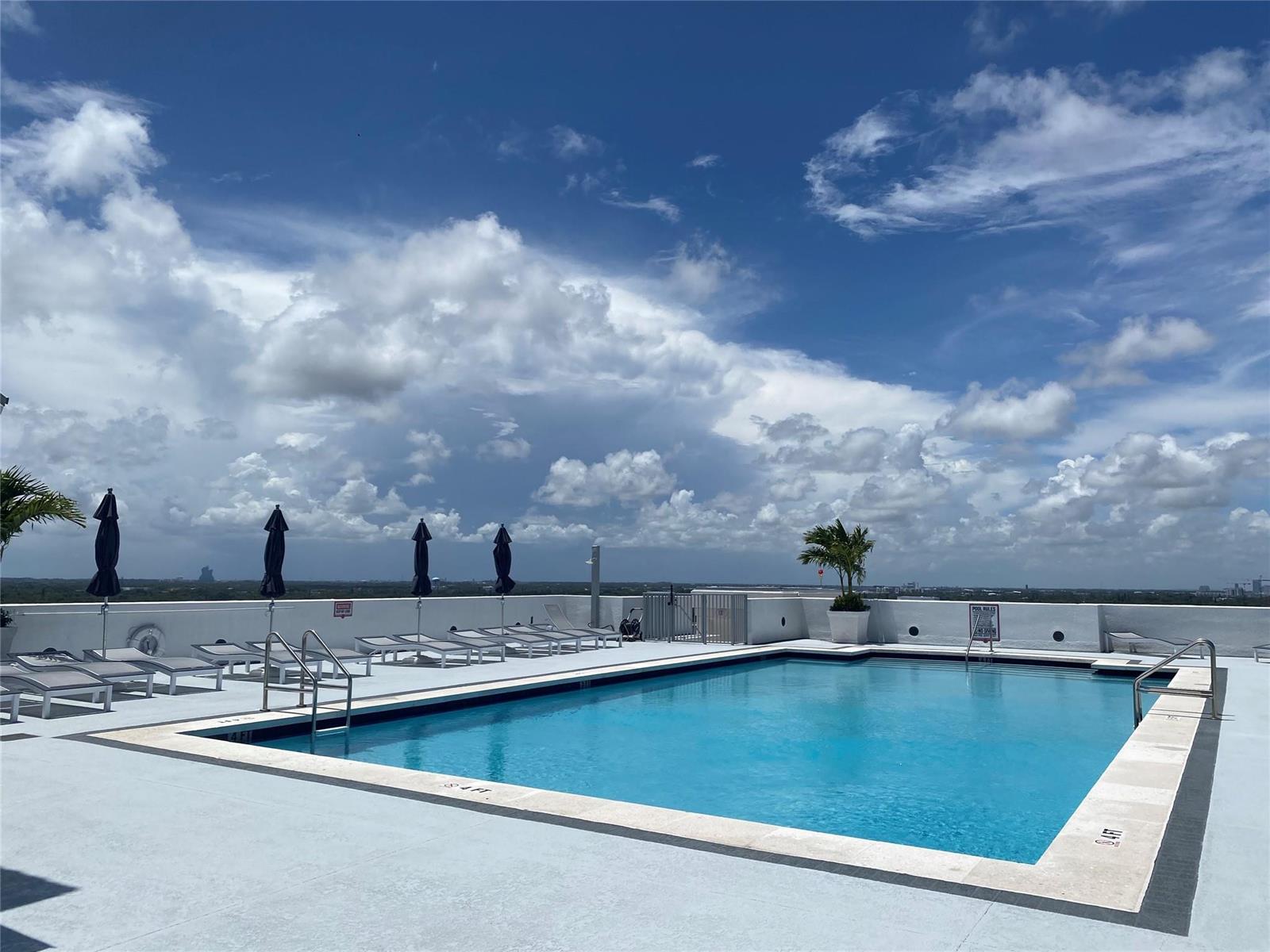 A rooftop swimming pool with lounge chairs and umbrellas under a cloudy blue sky — 1830 Radius Drive, Hollywood FL 33020, HOLLYWOOD, FL