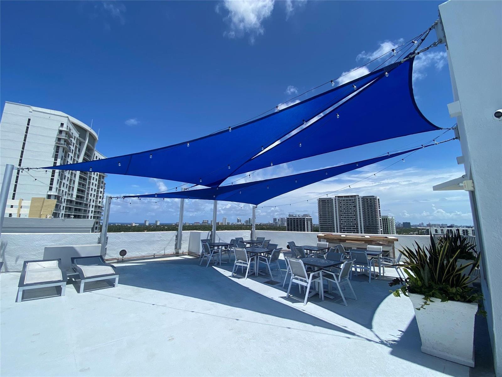 A rooftop terrace features blue shade sails, outdoor dining tables, and lounge chairs under a sky — 1830 Radius Drive, Hollywood FL 33020, HOLLYWOOD, FL