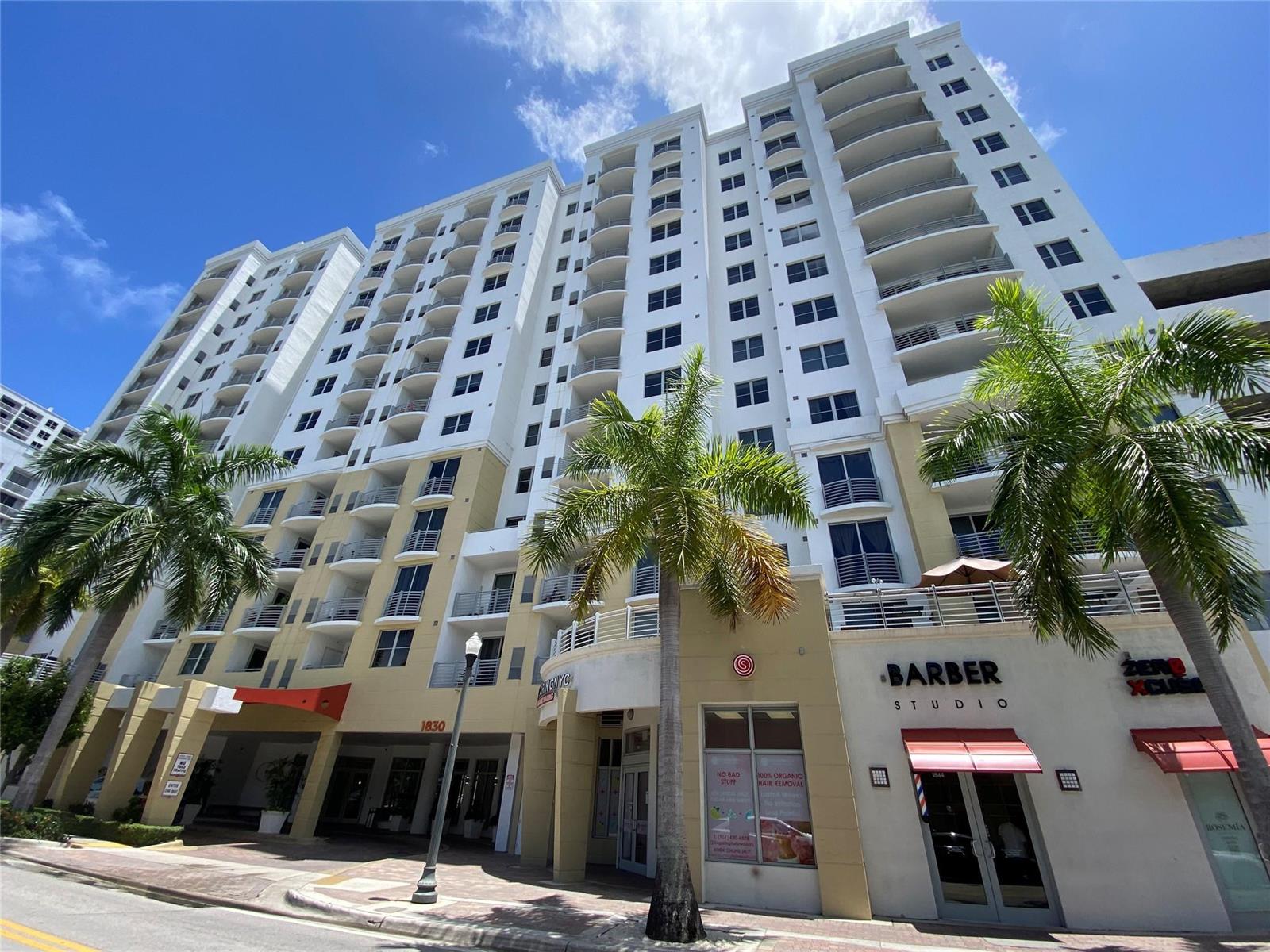 A low-angle view of a multi-story white apartment building with ground-floor retail storefronts and palm trees — 1830 Radius Drive, Hollywood FL 33020, HOLLYWOOD, FL
