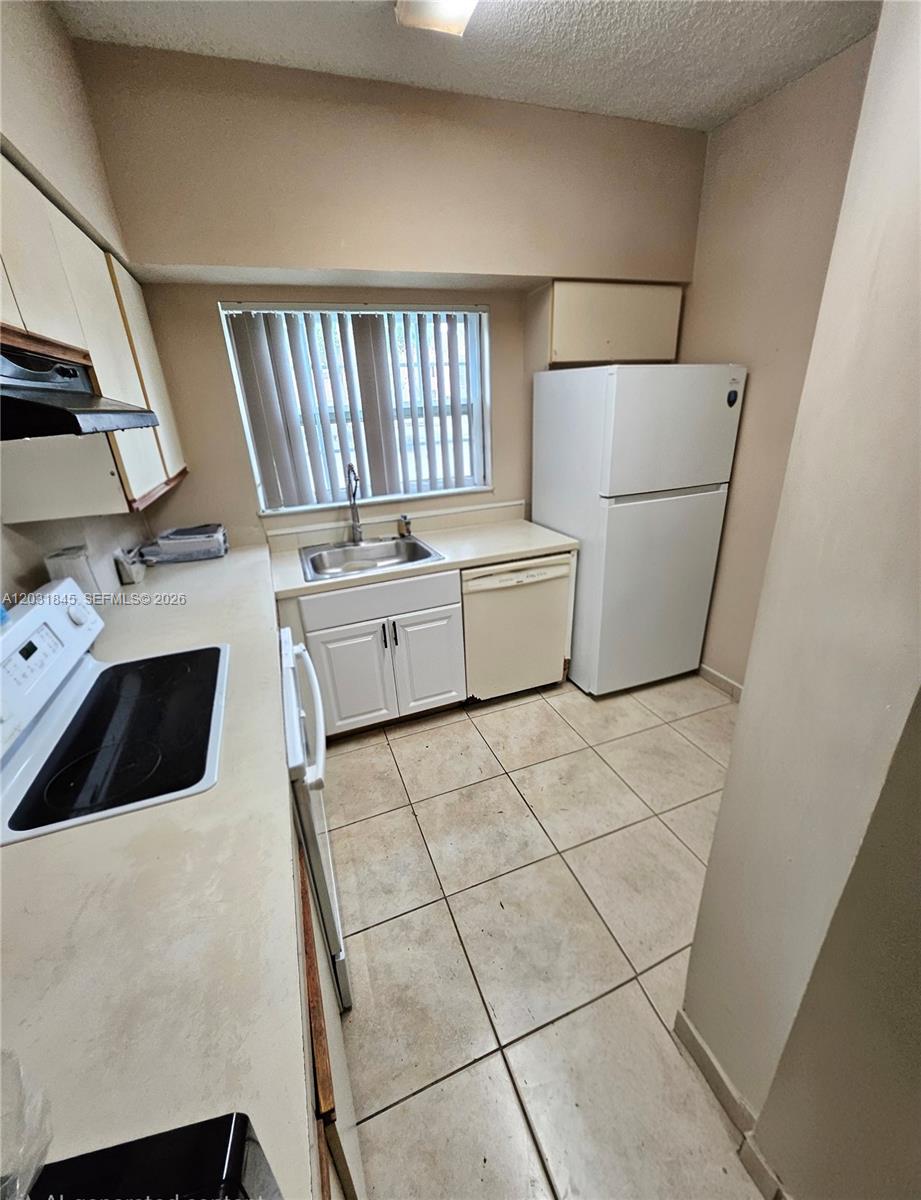 A small kitchen features white cabinets, a white refrigerator, a sink, and tiled flooring — 631 Cypress Lake Blvd # E17, Deerfield Beach FL 33064, DEERFIELD BEACH, FL