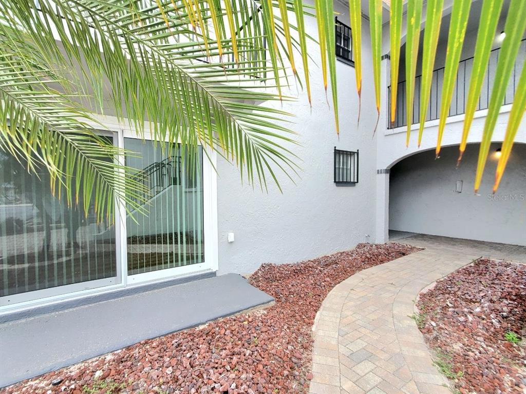 A paved walkway leads to a sliding glass door beside a gray stucco building exterior — 8151 BLIND PASS ROAD UNIT 3, ST PETE BEACH, FL