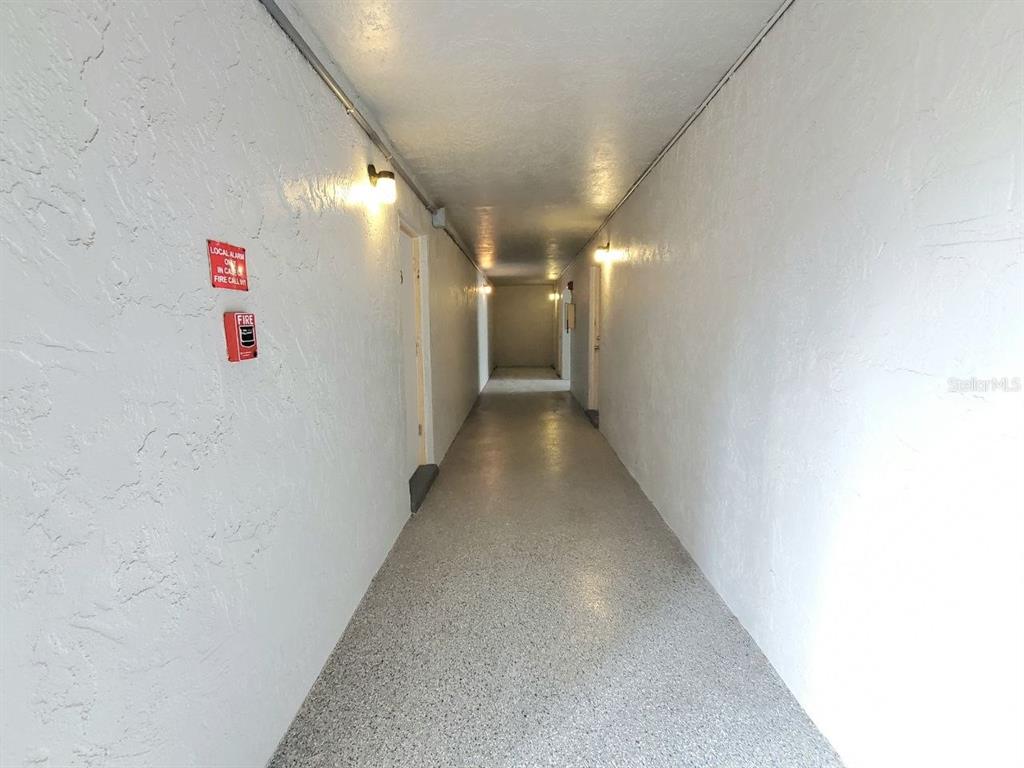 A long, narrow apartment hallway with white walls, textured surfaces, and light-colored speckled flooring — 8151 BLIND PASS ROAD UNIT 3, ST PETE BEACH, FL