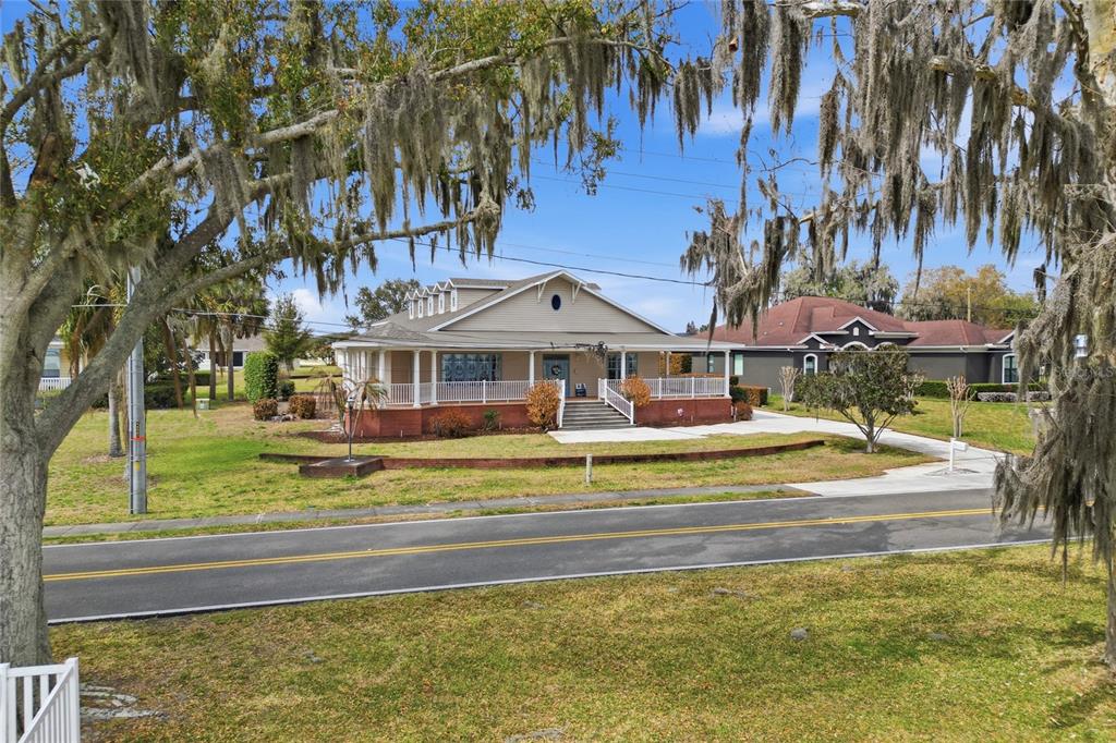 A tan house with a wrap-around porch and brick base sits behind a paved road — 2128 LAKE ARIANA BOULEVARD, AUBURNDALE, FL