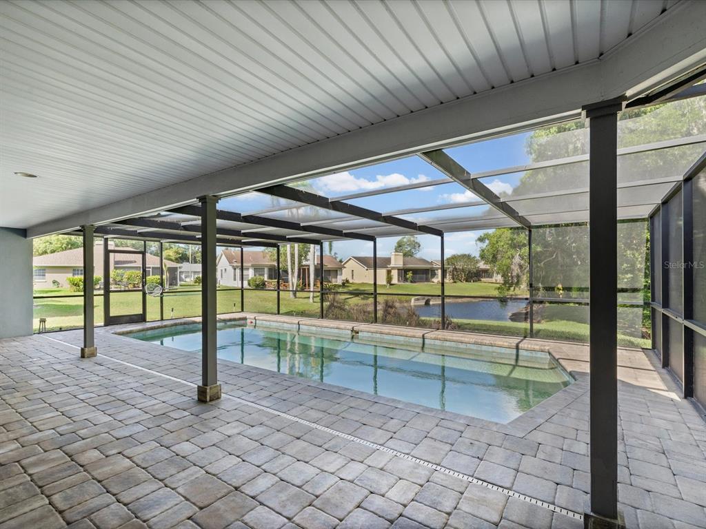 A screened-in patio features a rectangular swimming pool and paver flooring overlooking a pond — 3827 WATSON DRIVE, NEW PORT RICHEY, FL