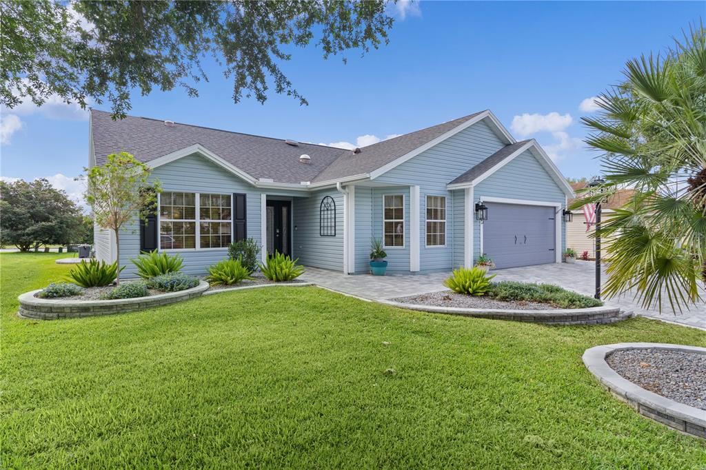 A light blue single-story house with a paved driveway, front lawn, and stone-bordered landscaping — 1795 HAGOOD LOOP, THE VILLAGES, FL