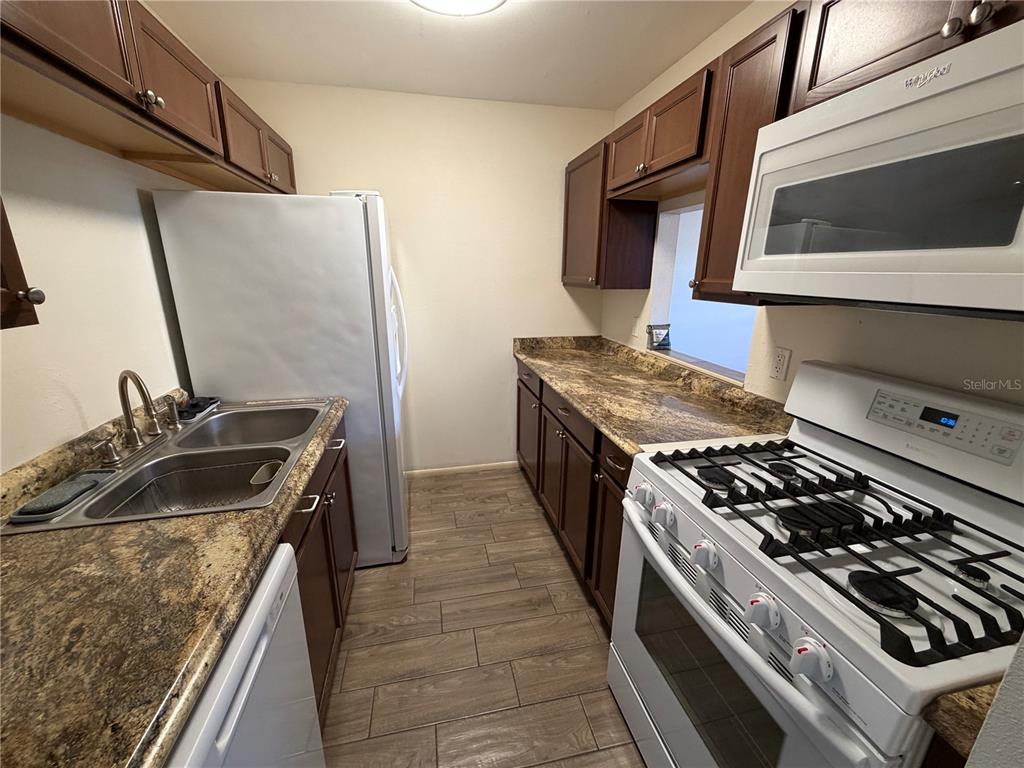 A galley kitchen features dark wood cabinets, granite countertops, and white appliances — 5967 TERRACE PARK DRIVE N UNIT 104, ST PETERSBURG, FL